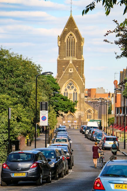 A street scene on Church Road in Barnes showing a row of parked cars along one side of the road, with a person walking and pushing a bicycle on the pavement. The foreground includes various vehicles such as compact and hatchback cars, some with visible registration plates, and a partially loaded moving van parked on the street. In the background, a large historic church with a tall, pointed steeple and arched windows dominates the scene, surrounded by green trees and other buildings. The sky is partly cloudy with patches of blue, and street lamps line the pavement. The image conveys a typical urban setting suitable for house removals or furniture transport, with visual cues like parked vehicles, loading activity, and the church providing context for moving or packing and moving services by Removal Van Barnes during a home relocation in Barnes.