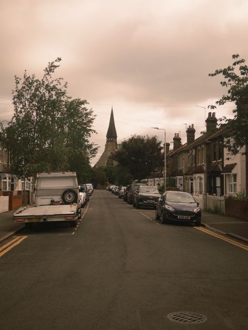 A narrow residential street on a cloudy day, showing parked vehicles including a white van with an open rear liftgate and various cars aligned along the pavement on both sides. The street is lined with terraced houses with brick facades, some with small front gardens, and chimneys visible on rooftops. In the background, a church spire rises above the treetops, indicating a historic area. The scene depicts a typical suburban setting suitable for house removals or relocating services, as provided by Removal Van Barnes, focusing on the logistics of furniture transport and loading process in an urban environment.