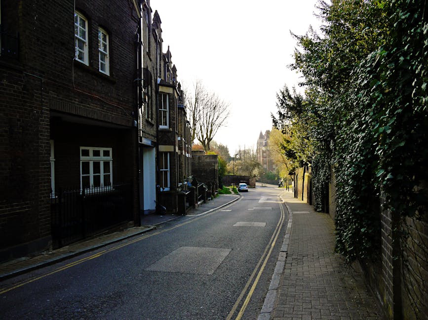 A street scene on Church Road in Barnes showing a row of parked cars along one side of the road, with a person walking and pushing a bicycle on the pavement. The foreground includes various vehicles such as compact and hatchback cars, some with visible registration plates, and a partially loaded moving van parked on the street. In the background, a large historic church with a tall, pointed steeple and arched windows dominates the scene, surrounded by green trees and other buildings. The sky is partly cloudy with patches of blue, and street lamps line the pavement. The image conveys a typical urban setting suitable for house removals or furniture transport, with visual cues like parked vehicles, loading activity, and the church providing context for moving or packing and moving services by Removal Van Barnes during a home relocation in Barnes.